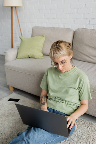 wytatuowana kobieta z grzywką i okularami za pomocą laptopa siedząca na dywanie obok smartfona z czystym ekranem i wygodną kanapą w nowoczesnym salonie  - Zdjęcie, obraz