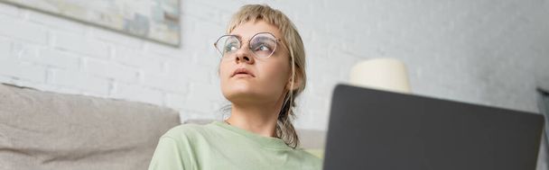 Düşük açılı sarışın, kısa saçlı, kâküllü ve gözlüklü bir kadının modern oturma odasındaki rahat kanepenin yanında otururken dizüstü bilgisayar kullanması, pankart, pankart.  - Fotoğraf, Görsel
