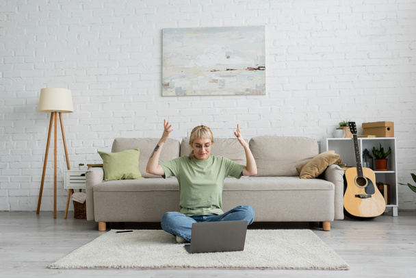 mujer tatuada emocional con flequillo y gafas usando el ordenador portátil mientras está sentado en la alfombra cerca de teléfono inteligente, cómodo sofá, guitarra y rack con plantas en la sala de estar moderna con paiting en la pared  - Foto, imagen