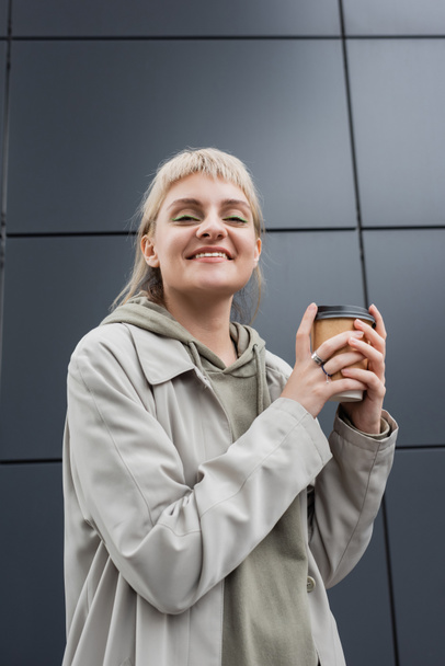 низкий угол зрения счастливой молодой женщины с блондинистыми волосами с челкой, стоящей в пальто и толстовке, держа бумажную чашку с кофе на вынос возле серого современного здания на улице, снаружи, городской жизни  - Фото, изображение