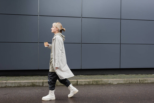 side view of young woman with blonde hair with bangs walking in coat, black leather pants, hoodie and boots while holding paper cup with takeaway coffee near grey modern building on street  - Photo, Image