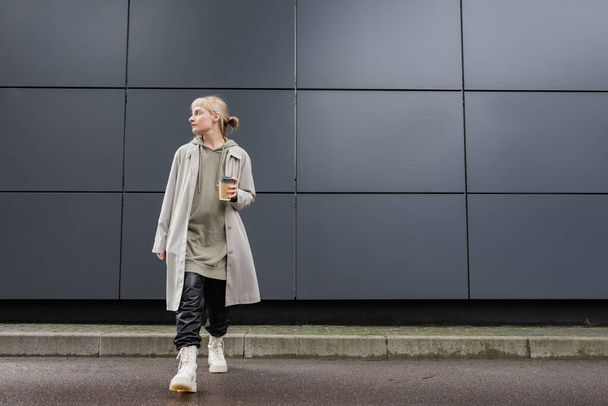 pełna długość młodej kobiety z blond włosami z grzywką chodzącej w płaszczu, czarnymi skórzanymi spodniami, bluzą z kapturem i butami trzymającej papierowy kubek z kawą na wynos w pobliżu szarego nowoczesnego budynku na ulicy  - Zdjęcie, obraz