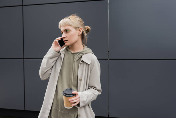 κομψή νεαρή γυναίκα με αφέλειες και ξανθά μαλλιά κρατώντας χάρτινο κύπελλο με καφέ για να πάει, ενώ μιλάμε για smartphone και στέκεται σε hoodie και παλτό κοντά γκρι σύγχρονο κτίριο σε αστικό δρόμο  - Φωτογραφία, εικόνα