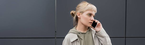 stylowa młoda kobieta z grzywką i blond włosami rozmawiająca na smartfonie i stojąca w bluzie i płaszczu w pobliżu szarego nowoczesnego budynku na ulicy miejskiej, baner, odwracając wzrok, miejski styl życia  - Zdjęcie, obraz