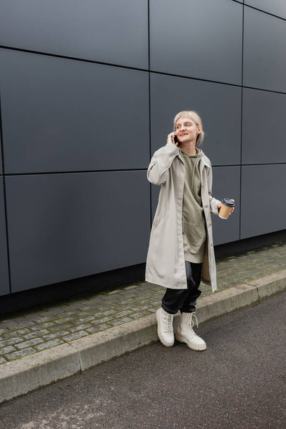 glückliche junge Frau mit Pony und blonden Haaren, die Pappbecher mit Coffee to go hält, während sie mit dem Smartphone spricht und in Kapuzenpulli und Mantel in der Nähe eines grauen modernen Gebäudes an der Stadtstraße steht  - Foto, Bild