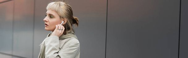jeune femme à la mode avec des franges tenant tasse en papier avec café à emporter tout en ajustant les écouteurs sans fil et debout dans une tenue à la mode près du bâtiment moderne gris sur la rue urbaine, bannière  - Photo, image