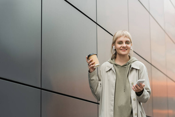 jeune femme souriante avec frange tenant tasse en papier avec café à emporter et debout dans une tenue à la mode et écouteurs sans fil tout en utilisant un smartphone près du bâtiment moderne gris dans la rue urbaine, regardez la caméra - Photo, image