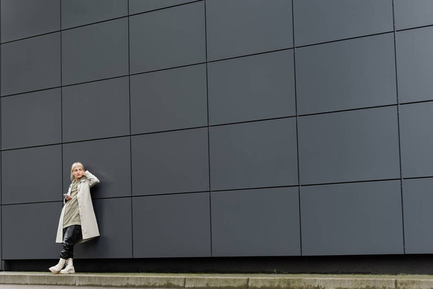 giovane donna con frangia in piedi in abiti eleganti e auricolari wireless durante l'utilizzo di smartphone vicino a grigio edificio moderno sulla strada urbana, cappotto con cappuccio, pantaloni e stivali in pelle nera, lunghezza intera - Foto, immagini
