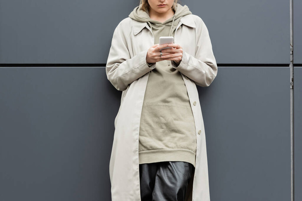 cropped view of fashionable young woman standing in trendy clothes and texting on smartphone near grey wall of modern building on urban street, coat with hoodie, partial view - Photo, Image