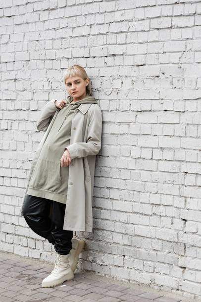 young woman with makeup, blonde hair, bangs, in stylish outfit, long hoodie, coat, black leather pants and beige boots standing near grey brick wall of modern building and looking away  - Photo, Image