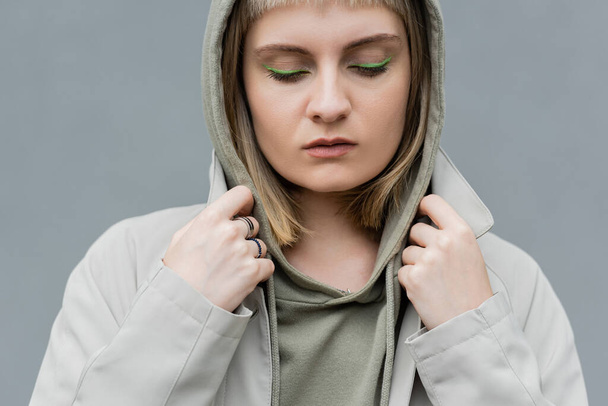 stylish and young woman with bangs, green eye shadows and blonde hair standing with hood on head and comfortable clothes while looking down isolated on grey background in studio, hoodie  - Photo, Image