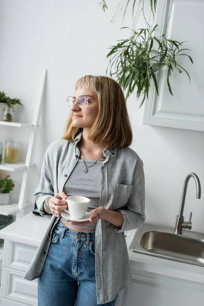 happy young woman with short hair and bangs, eyeglasses and tattoo holding cup of coffee while standing in casual grey clothes next to kitchen cabinets and plant in modern apartment  - Photo, Image