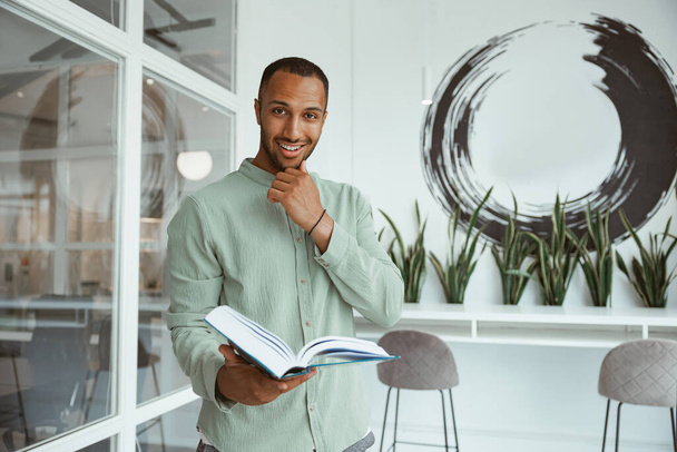 Smiling african businessman standing in modern office and holding book. Blurred background - Valokuva, kuva