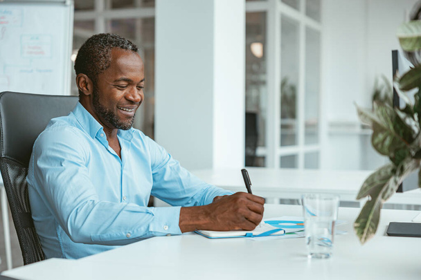 Mosolygó afrikai üzletember jegyzetel, miközben ül a modern irodában. Kiváló minőségű fénykép - Fotó, kép