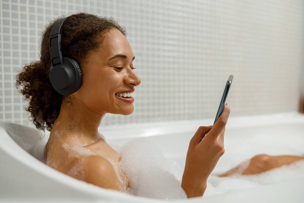 Afro american woman in headphones is using smartphone when taking a bath . High quality photo - Фото, зображення