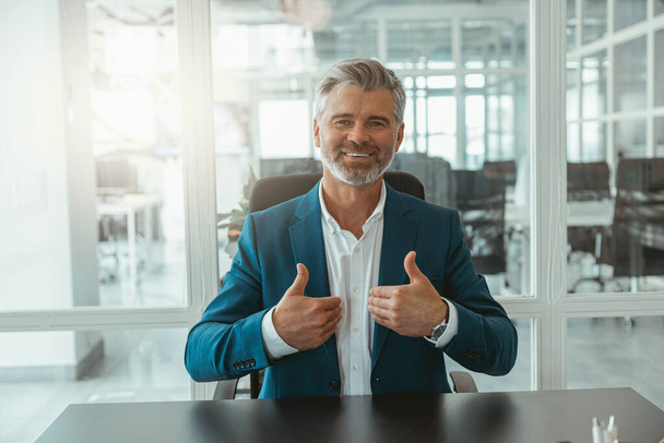 Smiling mature businessman showing raised fingers up while sitting in office. High quality photo - Foto, Imagem