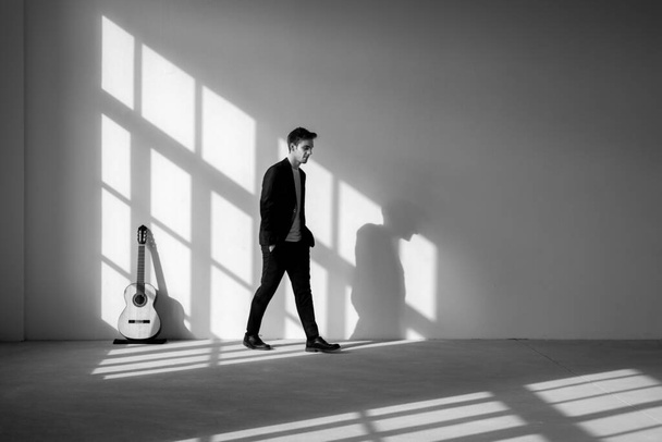 A standing boy is walking away from his guitar with his hands in his pockets. He is wearing a black suit. Interior of a renovated former factory. - Foto, Imagem