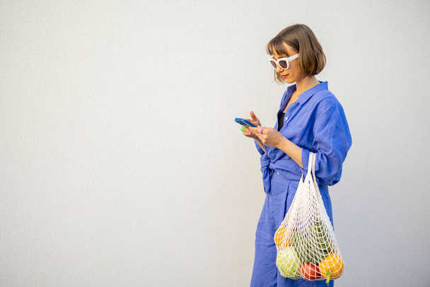 Woman in blue pajamas walks with mesh bag full of fresh fruits on white wall background outdoors. Concept of sustainability and modern lifestyle - Photo, Image
