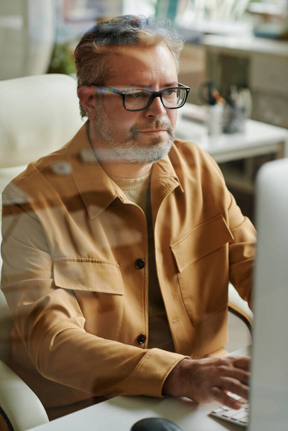 Moderno webdesigner maturo o programmatore seduto accanto a scrivanie davanti al monitor del computer e guardando lo schermo mentre lavora online - Foto, immagini