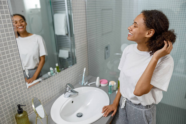 Smiling african woman standing in bathroom and looking in mirror. High quality photo - Photo, Image