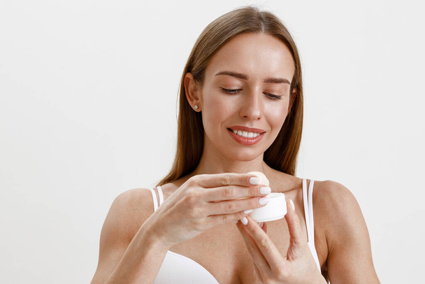 Vrouw draagt lingerie met gezichtscrème pot over witte studio achtergrond. Hoge kwaliteit foto - Foto, afbeelding