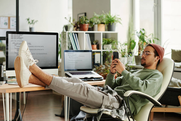 Junger IT-Ingenieur sitzt im Sessel am Arbeitsplatz mit Computermonitor und Laptop und SMS in der Pause - Foto, Bild