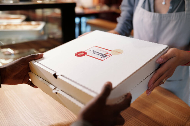 Close-up van de handen van caféeigenaar of serveerster die verpakte pizza aan bezorger over toonbank overhandigt terwijl hij hem helpt met het inpakken van zijn tas - Foto, afbeelding