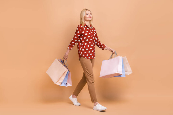 Full length profile portrait of positive lovely aged lady hold mall bags walking empty space isolated on beige color background. - Fotografie, Obrázek
