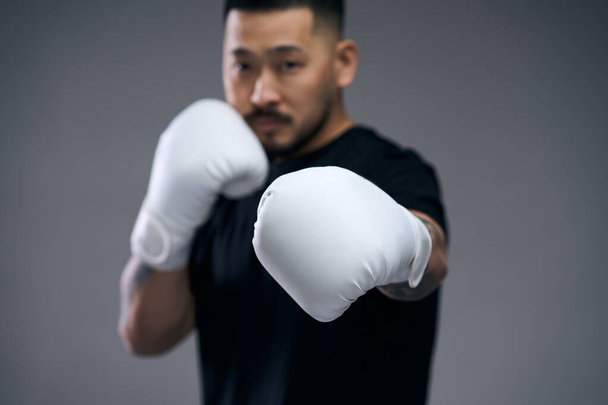 Portrait of asian young man in black t shirt hitting at camera in white boxing gloves isolated on grey background. Focus on hands  - Photo, Image
