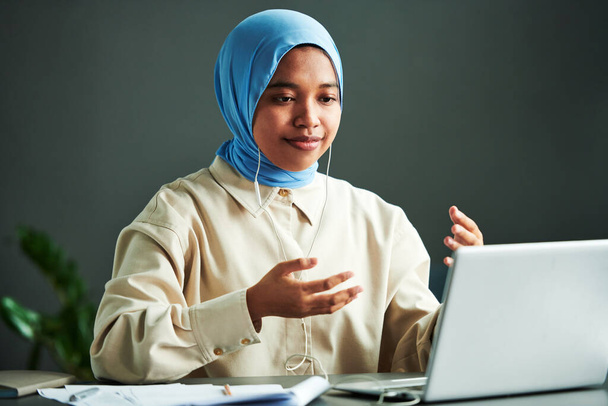 Mladý sebevědomý muslim žena v casualwear a modrý hidžáb vysvětlit něco, zatímco sedí před notebookem během on-line lekce - Fotografie, Obrázek