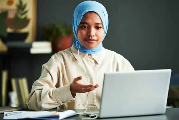 Mladá sebevědomá muslimka v hidžábu se účastní webináře, zatímco sedí u stolu před notebookem doma a něco vysvětluje - Fotografie, Obrázek