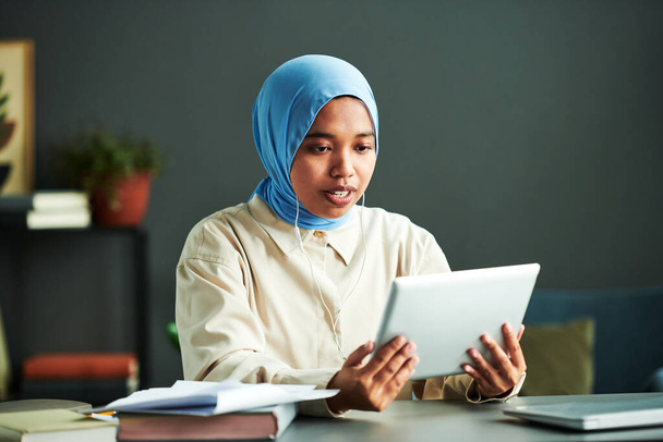 Mladá sebevědomá muslimka lektorka ve sluchátkách komunikující s online publikem při pohledu na obrazovku tabletu během lekce - Fotografie, Obrázek