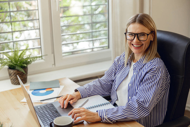 Attractive woman freelancer in eyeglasses working on laptop from home office. High quality photo - Foto, afbeelding