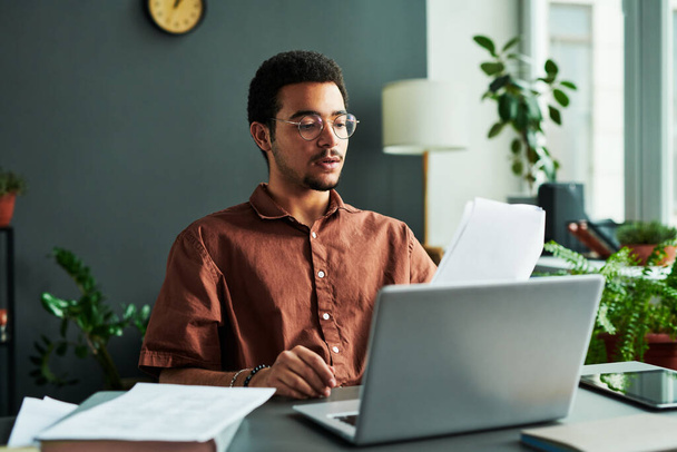 Jeune homme confiant avec des papiers assis par le lieu de travail devant un ordinateur portable et faire rapport ou expliquer de nouvelles informations à l'audience en ligne - Photo, image
