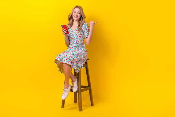 Full length photo of cheerful pretty lady wear flower print dress chatting gadget thumb empty space isolated yellow color background. - Fotoğraf, Görsel