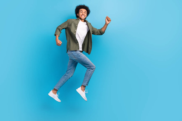 Full body photo of active young guy running wear khaki shirt denim jeans fast speed jumping positive isolated on blue color background. - Fotó, kép