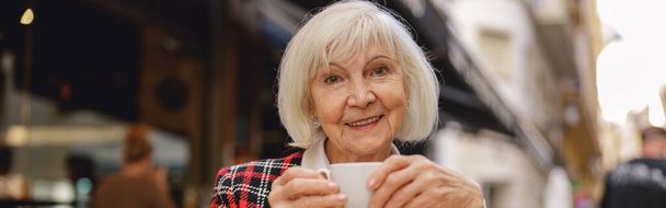 Senior señora urbana con elegante chaqueta expresando alegría mientras degustación taza de bebida en la acogedora cafetería - Foto, Imagen