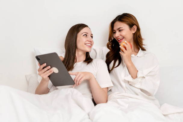若いアジアのカップルの女性は、タブレットを使用してインターネットを一緒にサーフィンし、毛布の下に横たわって、自宅でベッドルームで快適な枕にもたれている間にリンゴの果物を食べる. - 写真・画像