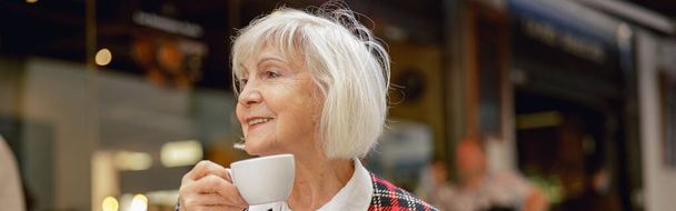 Femme âgée souriante portant une veste à carreaux à la mode regardant vers le côté tout en prenant un café dans le café de la ville - Photo, image
