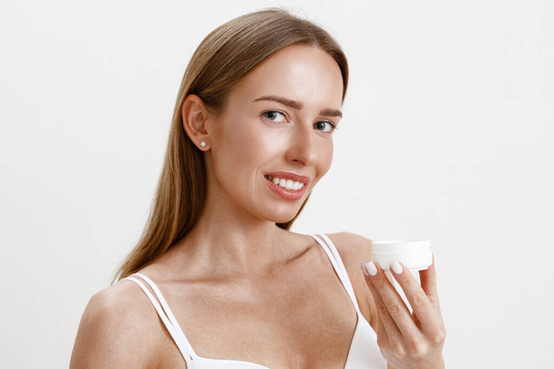 Slim smiling woman over white studio background wearing lingerie applying moisturizing cream - 写真・画像