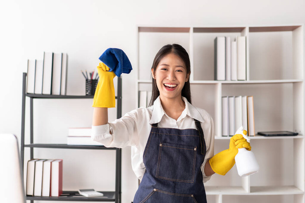 Huisvrouw in schort het dragen van handschoenen aan de hand van hygiëne spray fles en microvezel stof te vegen opruimen meubels, terwijl het werken met genieten van schoonmaken in het huis. - Foto, afbeelding