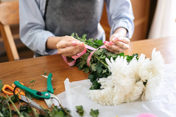 Female florist in apron using pink ribbon to tie for creating and making flower bouquet of white chrysanthemum in her flower shop for delivery to customer. - Foto, Bild