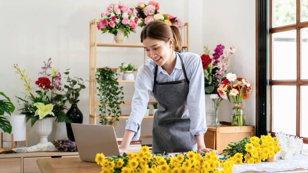 Vrouwelijke bloemist in schort met behulp van laptop om te werken en ontvangen orders van klanten tijdens het staan in haar bloemenwinkel te maken en het maken van bloemboeket met gele chrysant. - Foto, afbeelding