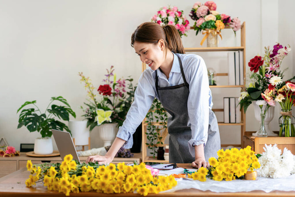 Γυναίκα ανθοπώλης με ποδιά που χρησιμοποιεί φορητό υπολογιστή για να εργάζεται και να λαμβάνει παραγγελίες πελατών ενώ στέκεται στο ανθοπωλείο της για να δημιουργήσει και να κάνει μπουκέτο λουλουδιών με κίτρινο χρυσάνθεμα. - Φωτογραφία, εικόνα