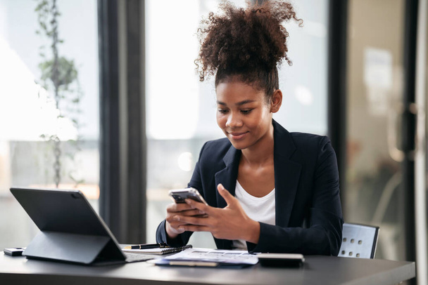 アフリカ系アメリカ人のビジネス女性がスマートフォンで新しいビジネスの会計情報を読み、現代のワークスペースで書類作業をしながらプロジェクトの戦略を考える. - 写真・画像