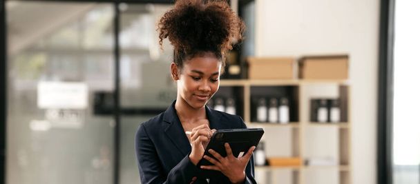 アフリカ系アメリカ人のビジネスウーマンは、タブレットを使用して新しいビジネスの会計情報を読み、現代のワークスペースでプロジェクトの作業と思考戦略に立ってメモを取ります. - 写真・画像