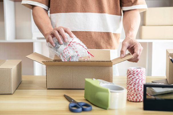 Mężczyzna przedsiębiorca pakuje produkt do tekturowych pudełek w celu przygotowania się do wysłania klienta podczas pracy online dostawy zakupów w biurze. - Zdjęcie, obraz