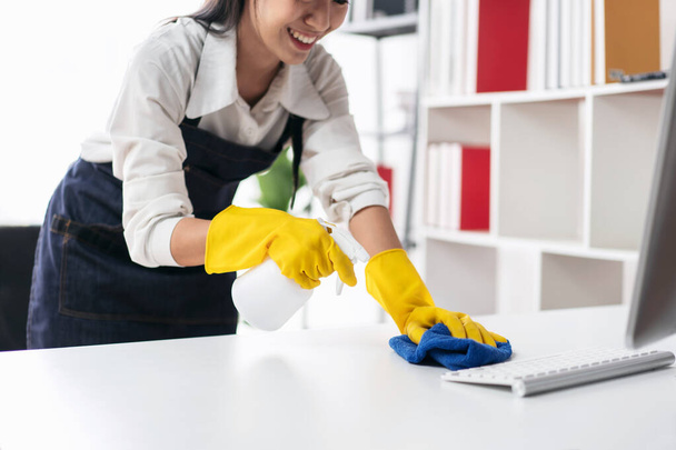 手袋を着用したエプロンの主婦は、コンピュータ上で衛生スプレーを噴霧し、家の中で家具を掃除しながらテーブルを拭くためにマイクロファイバーファブリックを使用しています。. - 写真・画像