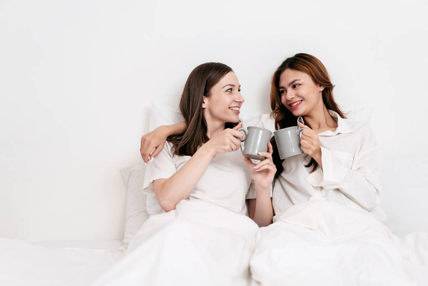 Young asian couple woman clinking cup and drinking chocolate cup while embracing and lying under blanket and leaning on comfortable pillows on the bed in bedroom at home. - Φωτογραφία, εικόνα