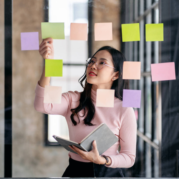 アイデアを共有する事業計画、ガラスの壁にメモを読み、オフィスでの事業計画について分析することを考えるビジネス女性. - 写真・画像